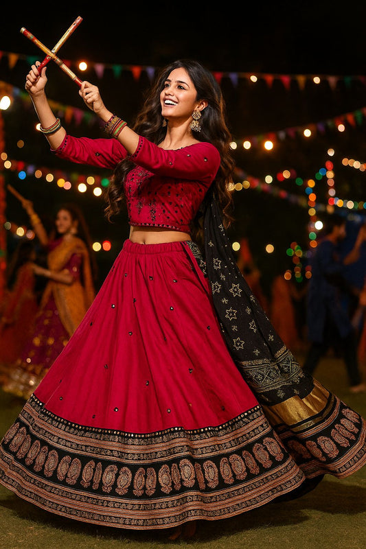 Woman in a red traditional outfit with decorative patterns, holding sticks, against a festive background with lights.