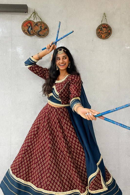 Woman in traditional maroon and blue outfit holding blue sticks against a light gray wall.